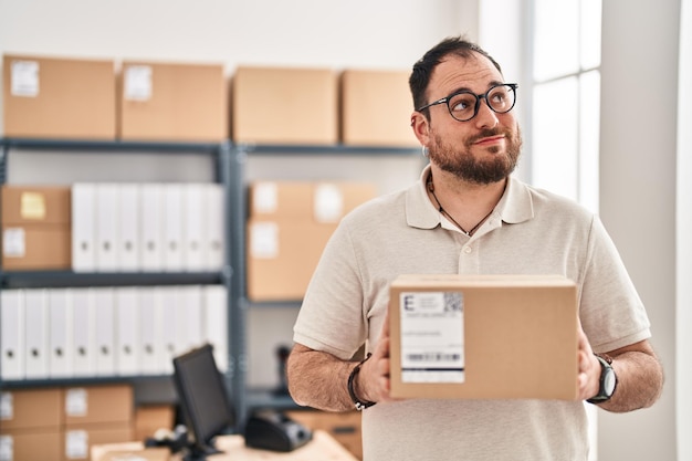 Hombre hispano de talla grande con barba que trabaja en el comercio electrónico de pequeñas empresas sonriendo mirando hacia un lado y mirando hacia otro lado pensando.