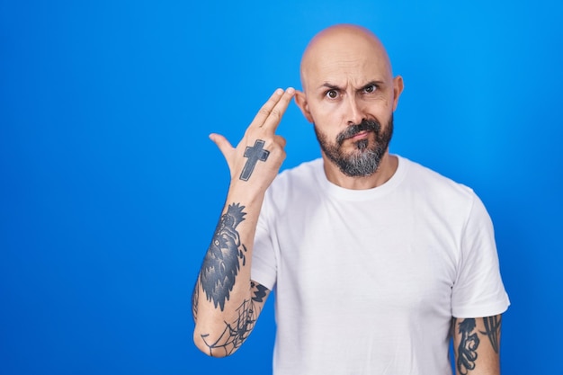 Hombre hispano con tatuajes de pie sobre fondo azul disparándose y matándose a sí mismo apuntando con la mano y los dedos a la cabeza como gesto de suicidio con pistola