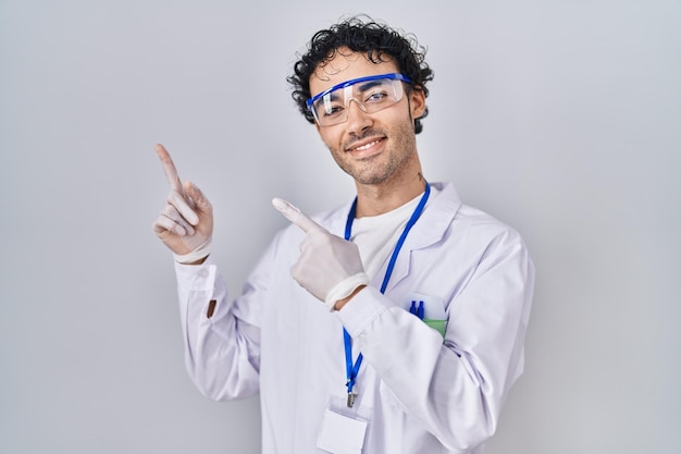 Hombre hispano trabajando en un laboratorio científico sonriendo y mirando la cámara apuntando con dos manos y dedos hacia un lado