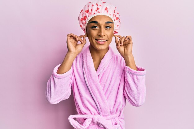 Hombre hispano vestido con maquillaje con gorro de toalla de ducha y albornoz sonriendo tirando de las orejas con los dedos gesto divertido problema de audición