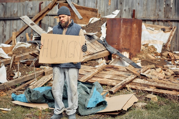 Un hombre sin hogar cerca de las ruinas con un cartel hambriento, ayudando a las personas pobres y hambrientas durante la epidemia.