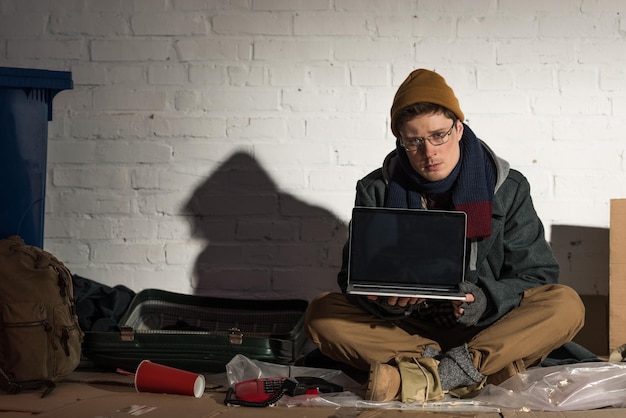 Hombre sin hogar serio que usa una computadora portátil mientras está sentado junto a una pared de ladrillos en un basurero