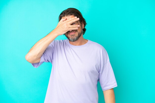 Hombre holandés mayor aislado de fondo azul cubriendo los ojos con las manos y sonriendo