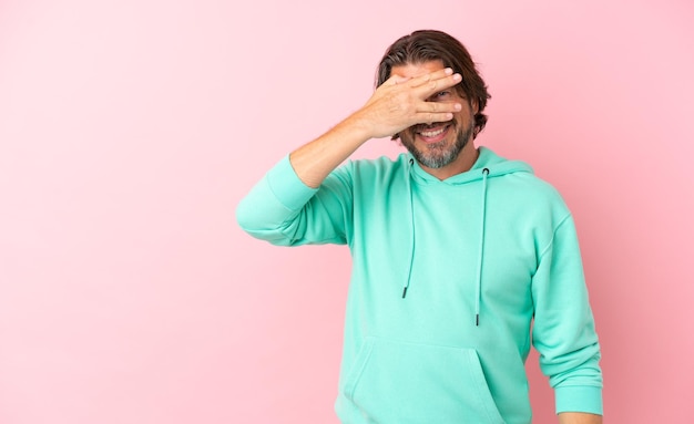 Hombre holandés mayor aislado de fondo rosa cubriendo los ojos con las manos y sonriendo