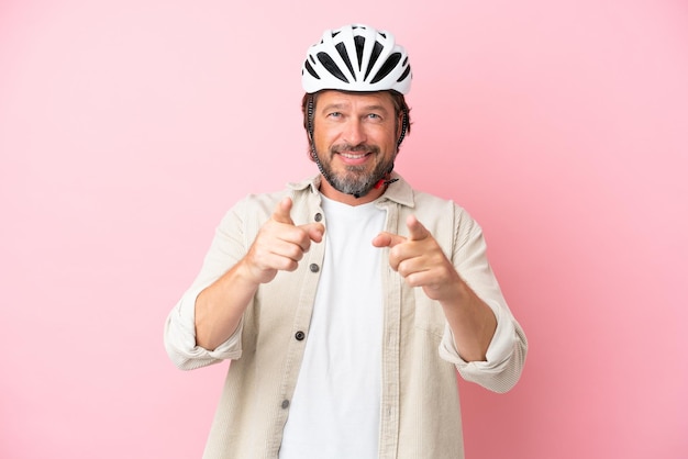 Hombre holandés mayor con casco de bicicleta aislado de fondo rosa apuntando al frente y sonriendo