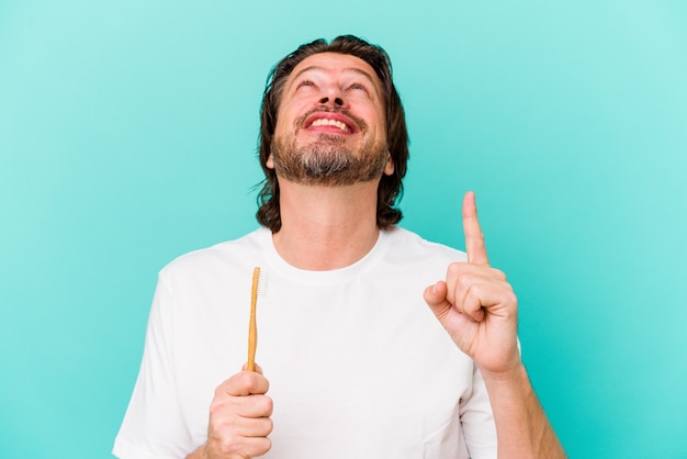 Hombre holandés de mediana edad sosteniendo un cepillo de dientes aislado en la pared azul apuntando hacia arriba con la boca abierta.