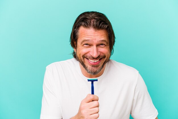 Hombre holandés de mediana edad sosteniendo una hoja de afeitar aislada en la pared azul riendo y divirtiéndose