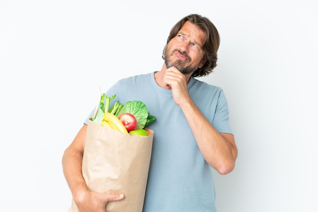 Hombre holandés senior sosteniendo la bolsa de la compra sobre fondo aislado teniendo dudas y pensando