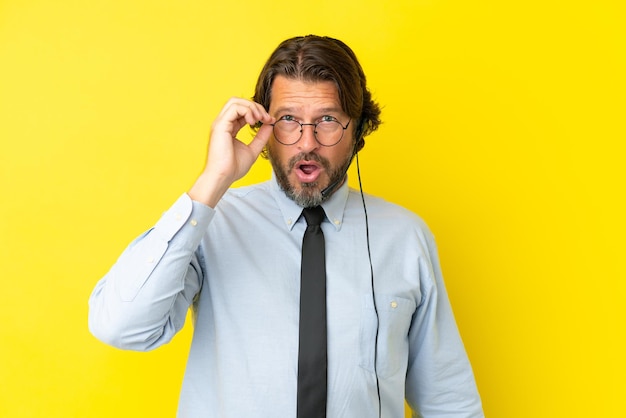 Hombre holandés telemarketer trabajando con un auricular aislado sobre fondo amarillo con gafas y sorprendido