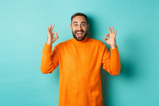 Hombre impresionado alabando algo impresionante, mostrando signos de estar bien y mirando asombrado, de pie con un suéter naranja sobre fondo azul claro.