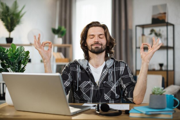 Foto hombre independiente con anteojos sentado en la mesa con los ojos cerrados y aliviando el estrés mediante la meditación