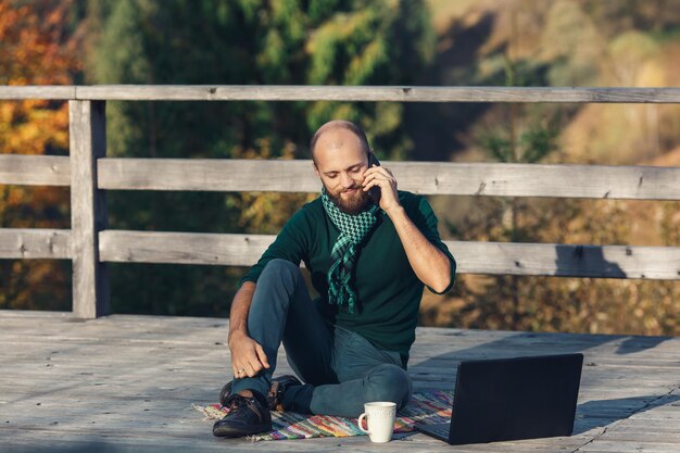 Hombre independiente barbudo sentado en la terraza, trabajando en una laptop y hablando por teléfono.