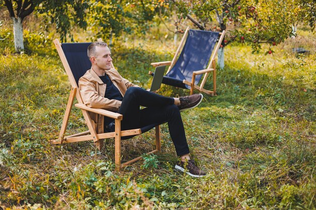 Hombre independiente con computadora portátil trabajando en el jardín sentado en una silla de madera al aire libre Un joven bloguero trabaja en una computadora en un parque público Lugar de trabajo trabajo remoto