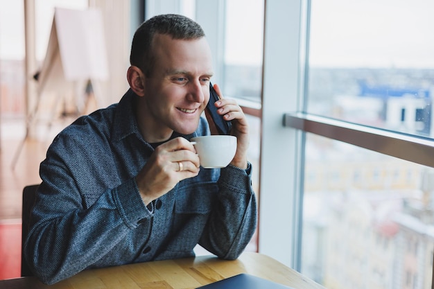 Hombre independiente europeo revisando la notificación en el teléfono inteligente sobre transacciones de dinero haciendo negocios en línea trabajando de forma remota mientras está en un café y bebiendo café