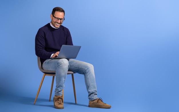 Hombre independiente feliz trabajando en un proyecto en una computadora portátil mientras está sentado en una silla contra un fondo azul