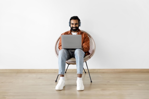 Hombre independiente indio que usa una computadora portátil con auriculares sobre una pared blanca