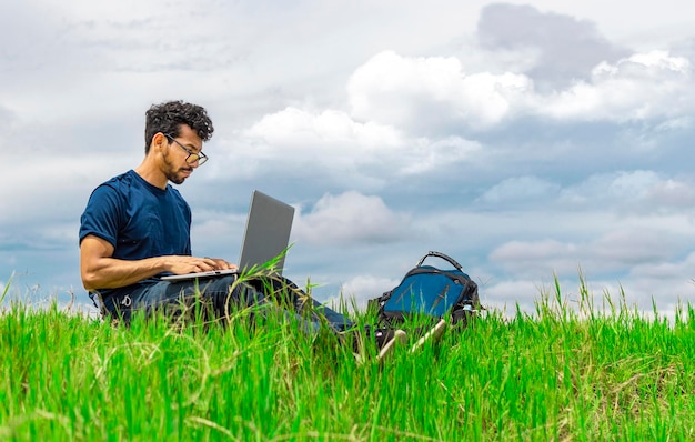 Hombre independiente sentado con una computadora portátil y una mochila en el campo Hombre sentado en el campo verde trabajando desde su computadora portátil Concepto de hombre independiente trabajando desde el campo