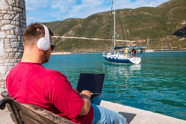 Hombre independiente trabajando en una computadora portátil en el banco del puerto de la ciudad en verano