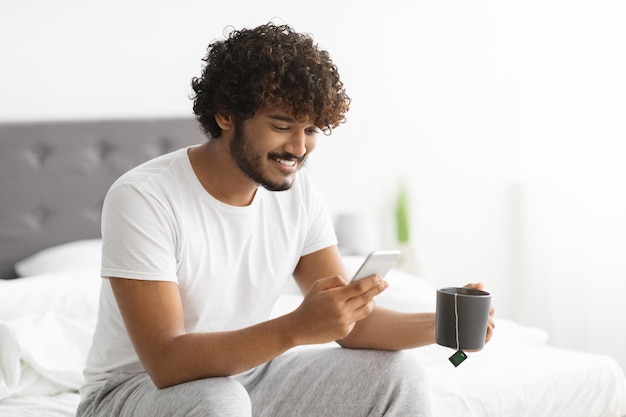 Hombre indio alegre bebiendo té y usando un teléfono inteligente