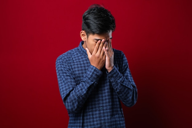 Hombre indio con bigote vistiendo camisa casual con expresión triste cubriendo el rostro con las manos mientras llora. concepto de depresión sobre fondo rojo