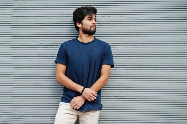 Hombre indio casual elegante usa camiseta azul posando contra la pared gris.