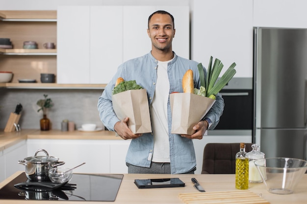 Hombre indio en la cocina sosteniendo dos bolsas de papel llenas de verduras frescas y saludables