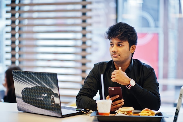 Hombre indio elegante sentado en el café de comida rápida contra su computadora portátil con teléfono móvil a mano.