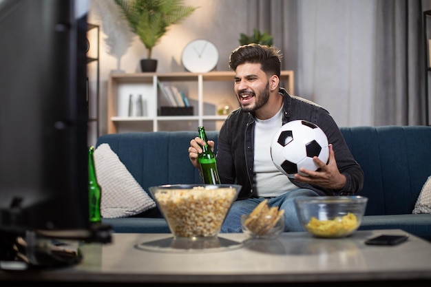 Hombre indio emocionado con papas fritas y cerveza viendo fútbol en la televisión