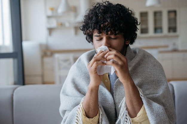 Hombre indio enfermo con secreción nasal sosteniendo servilletas de papel cerca de la cara sentado en casa Alergia al virus de la gripe