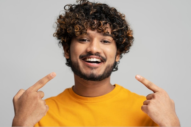 Hombre indio feliz y confiado con una sonrisa dentuda apuntando con los dedos en la boca mirando a la cámara