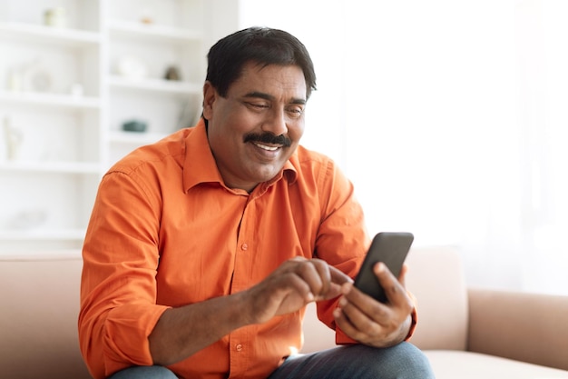 Hombre indio feliz usando el teléfono sentado en el sofá en casa