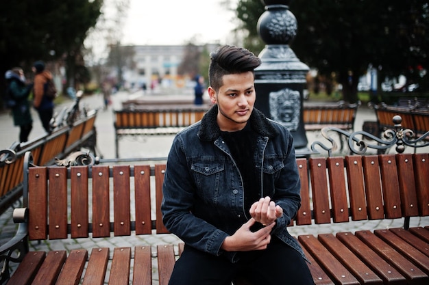 Hombre indio guapo y de moda en chaqueta de jeans negros posó al aire libre sentado en un banco