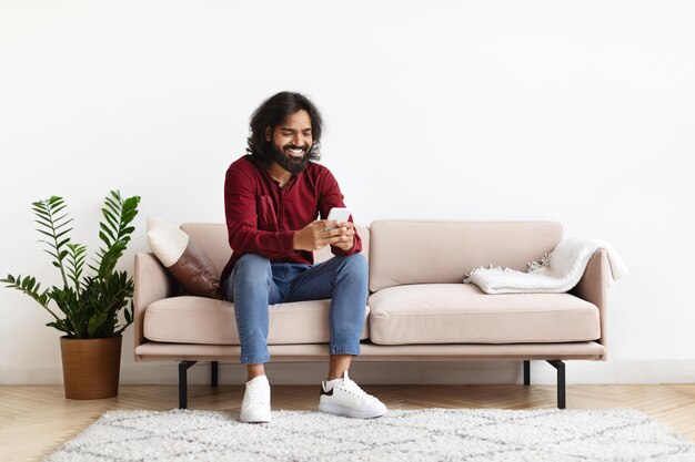 Hombre indio guapo usando teléfono inteligente mientras se relaja en el sofá