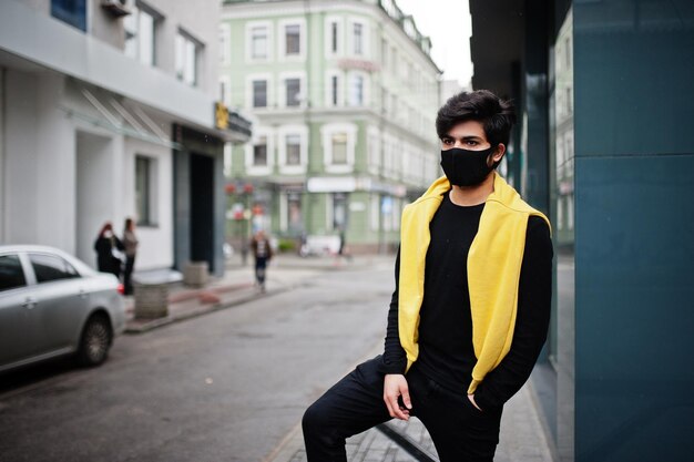 Hombre indio joven hipster urbano en una sudadera amarilla de moda Chico fresco del sur de Asia usa sudadera con capucha y máscara de protección de cara negra durante la nueva normalidad