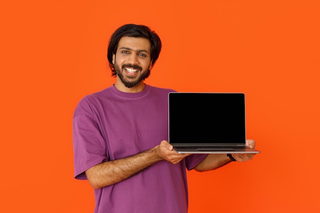 Hombre indio positivo que muestra una computadora portátil con pantalla en blanco en naranja