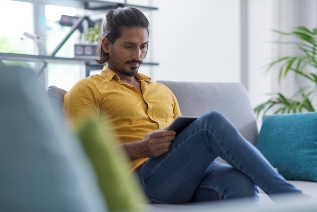 Hombre indio sentado en el sofá en casa y conectándose en línea usando una tableta digital