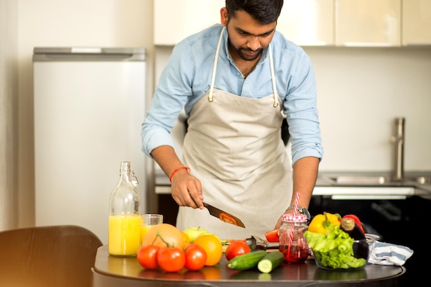 Hombre indio sonriente alegre hermoso que cocina la ensalada de verano para la cena, cortando el tomate en la tabla de cortar, preparando la cena para la esposa en casa en la cocina. Comida sana, concepto de cocina.