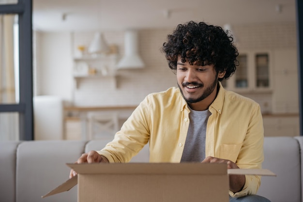 Hombre indio sonriente desempacando caja con presente sentado en casa Concepto de entrega de compras en línea