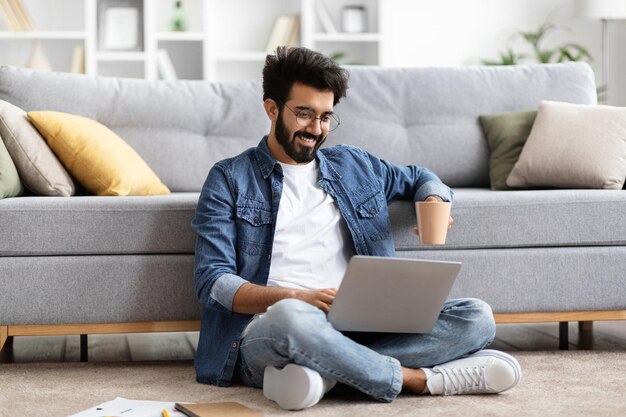 Hombre indio sonriente usando una computadora portátil mientras está sentado con las piernas cruzadas en el suelo de su casa