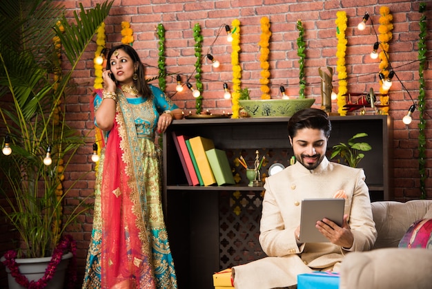 Hombre indio con tableta o tableta con computadora con pantalla táctil y esposa iluminando diyas en el fondo el día del festival de Diwali