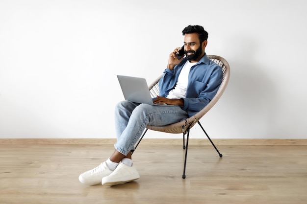Hombre indio teniendo una conversación telefónica y usando una computadora portátil sentado en el interior