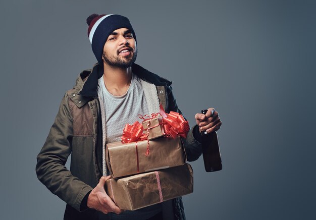 Un hombre indio vestido con una chaqueta cálida y un sombrero sostiene una botella con cerveza artesanal y un regalo de Navidad sobre un fondo gris.