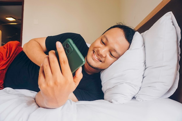 Hombre indonesio sonriente con teléfono cuando en la cama está acostado de lado y relajado