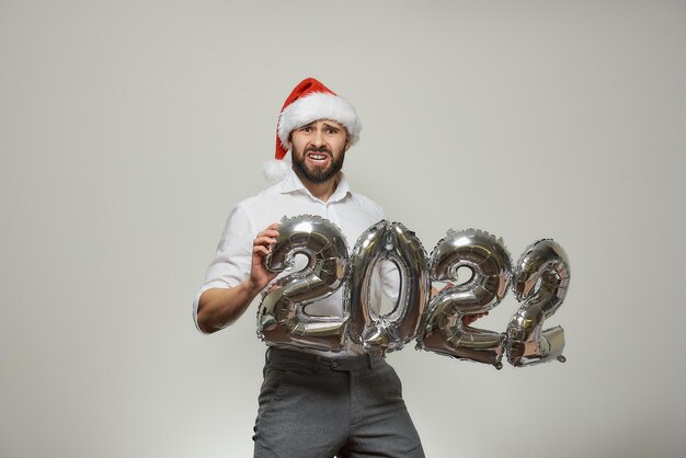 Un hombre infeliz con un gorro de Papá Noel de terciopelo rojo sostiene globos plateados con la forma de 2022. Un hombre con barba en una fiesta de año nuevo.