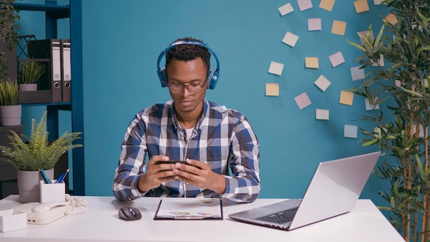 Hombre infeliz jugando juegos móviles en smartphone y perdiendo, disfrutando de la competencia en línea con auriculares. Jugador frustrado usando teléfono móvil y auriculares en el escritorio, sintiéndose decepcionado.