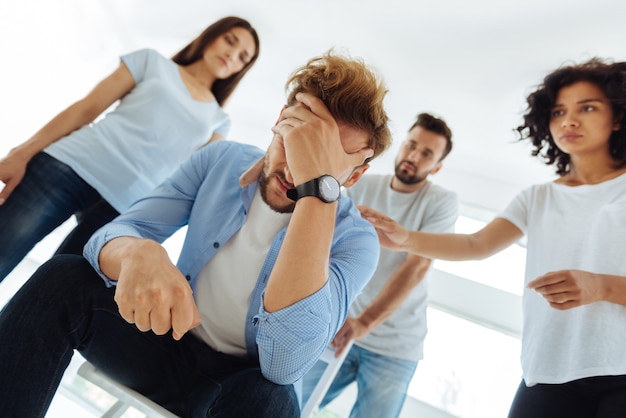 Hombre infeliz malhumorado deprimido sosteniendo su frente y pensando en cómo hacer frente a los problemas mientras está rodeado por los miembros de su grupo