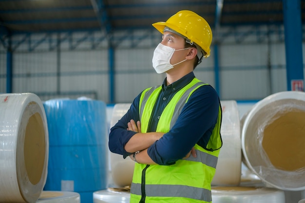 Un hombre de ingeniería con máscara médica, casco protector que trabaja en la fábrica de almacén