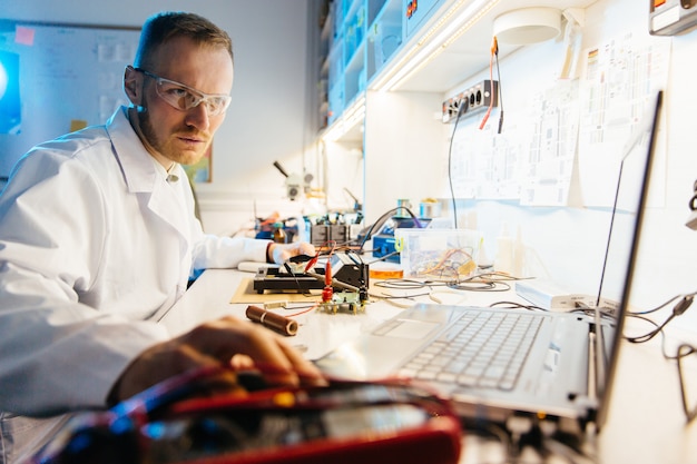 Hombre ingeniero en bata blanca usando laptop para trabajar en laboratorio electrónico