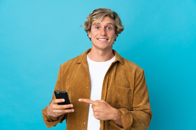 Hombre inglés sobre pared azul aislada mediante teléfono móvil y apuntando