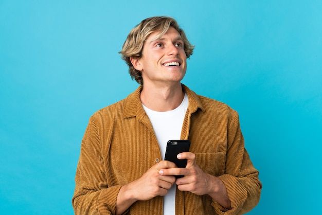 Hombre inglés sobre pared azul aislada con teléfono móvil y mirando hacia arriba